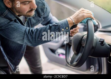 Young handsome bearded man, cleaning his modern futuristic car interior, car steering wheel with microfiber clothes. Car detailing and valeting Stock Photo
