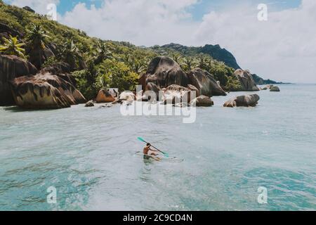 "La digue" island in Seychelles. Silver beach with granitic stone, and jungle. Man enjoying vacations on the beach and having fun with kayak. Aerial v Stock Photo