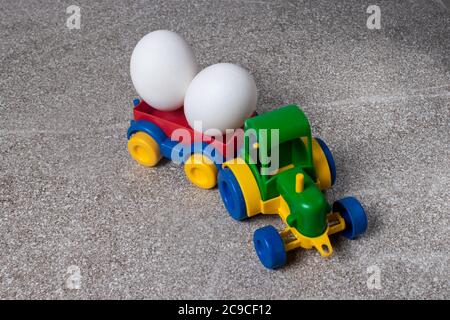 A toy tractor carries two chicken eggs. Plastic and natural food. Stock Photo