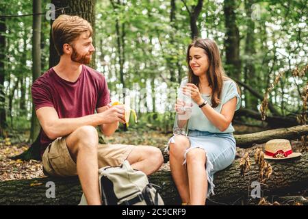 young couple sat to rest during forest walk along path to fallen tree, people drink water and eat banana fruit. Tourists break for snacks and Stock Photo