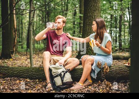 young couple sat to rest during forest walk along path to fallen tree, people drink water and eat banana fruit. Tourists break for snacks and Stock Photo