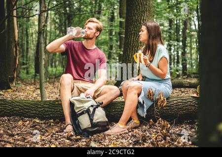People eat banana and drink water from plastic bottle while sitting on log in wood. Couple hikers take break for food and drink in forest on fallen Stock Photo