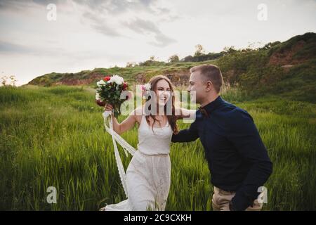 Outdoor wedding ceremony, stylish happy smiling groom and bride are laughing and looking at each other in the green field Stock Photo
