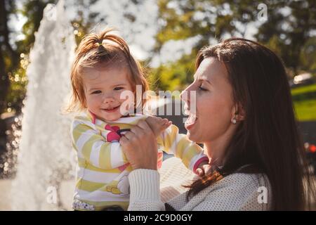 Close up portrait of beautiful small baby, blond little girl in colourful pullover in mothers hands in park outdoors Stock Photo
