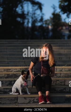 Woman sits next to her dog, natural sunlight, urban scene. Lifestyle with pets in town, smooth fox terrier breed Stock Photo