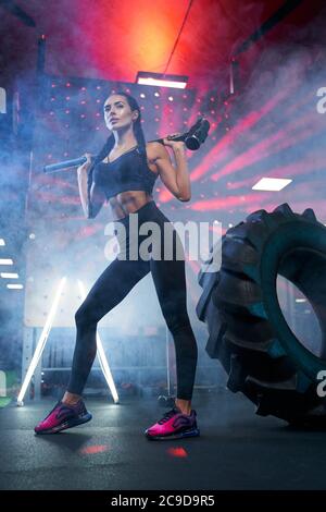 From below view of muscular woman in sportswear holding sledgehammer on shoulders near tire wheel, smoky atmosphere. Portrait of fit female bodybuilder posing in gym, having rest after hard training. Stock Photo