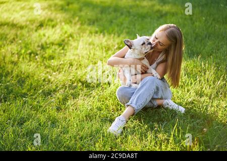 Lovely french bulldog kissing attractive human female friend. From above view of stunning caucasian woman enjoying summer sunset, hugging cute dog in city park. Human and animal friendship. Stock Photo