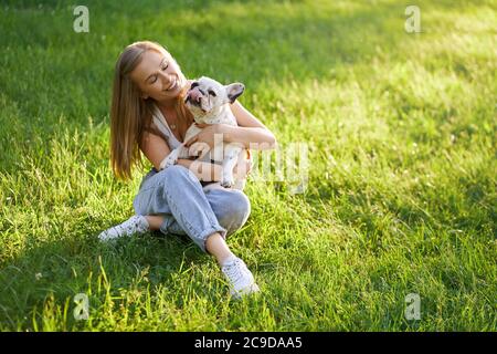 Top view of young happy woman sitting on grass with friendly french bulldog. Gorgeous caucasian smiling girl enjoying summer sunset, holding dog on knees and showing love and care in city park. Stock Photo