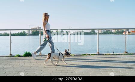Side view of young happy woman in casual outfit walking with male french bulldog on leash near big lake. Stunning caucasian girl wearing sunglasses enjoying summer sunset with pet in city park. Stock Photo