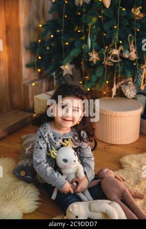 Christmas portrait of happy smiling beautiful little girl sitting on floor with presents under the christmas tree. Winter holiday Xmas and New Year Stock Photo