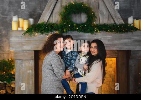 Christmas family portrait of happy smiling mothers hugging their children near to fireplace decorated with fir and garland at home. Winter holiday Stock Photo