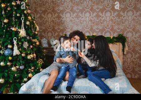 Christmas family portrait of happy smiling women sitting on bed hugging and playing with their small children near to christmas tree at home. Winter Stock Photo