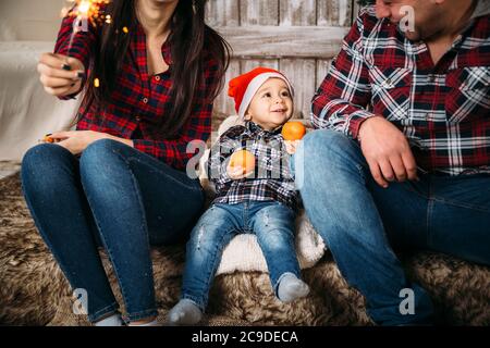 Christmas family portrait of little kid in red santa hat with oranges in hands sitting between parents. Winter holiday Xmas and New Year concept Stock Photo