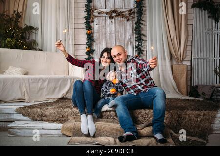 Christmas family portrait of young happy smiling parents with little kid in red santa hat holding sparklers. Winter holiday Xmas and New Year concept Stock Photo