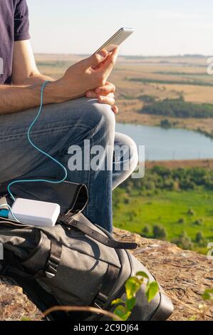 Man on a hike uses smartphone while charging from the power bank on the rock at dawn. Healthy lifestyle and communication Stock Photo