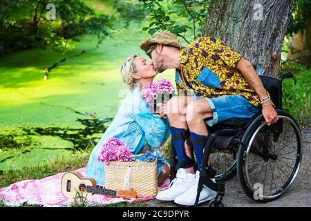 man in wheelchair kissing blond woman sitting by a river Stock Photo