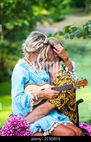 blond woman with guitar sitting on lap of man in wheelchair outdoors and kissing him Stock Photo