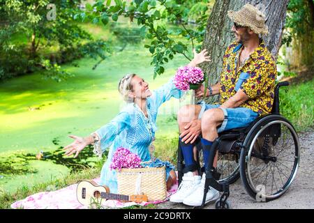 blond woman sitting by river spreading her arms toward a man in wheelchair Stock Photo