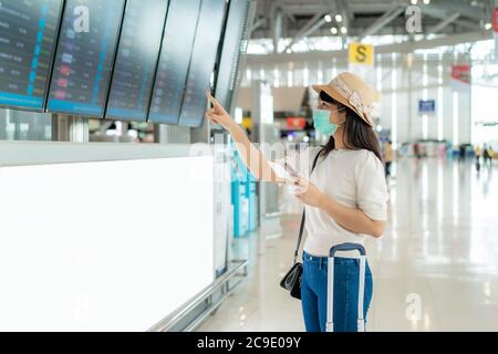 Asian woman tourist wearing face mask checking flight from arrival departure board in airport terminal for new normal travel concept. Stock Photo