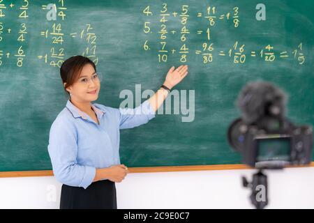 Asian primary teacher tutor explaining math in blackboard giving remote school class online lesson teaching looking at camera in classroom by e-learni Stock Photo