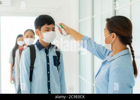 Asian woman teacher using thermometer temperature screening student for fever against the spread of COVID-19 while student coming back to school, New Stock Photo