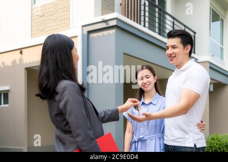 Asian happy smile young couple take keys new big house from real estate agent or realtor in front of their house after signing contract agreement, con Stock Photo