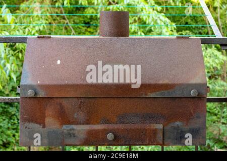 Old rusty large size barbecue grill with ash tray and chimney hanging on iron bars with trees in the background. Stock Photo