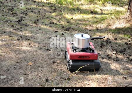 An aluminum saucepan with a closed lid stands on a camping gas stove on the sand in the forest. Karakansky Bor, Novosibirsk Region. Stock Photo