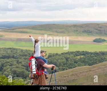 Daughter sits on father's shoulders with hands up. The concept of fatherhood and an active lifestyle with children. Stock Photo