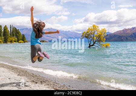 New Zealand travel happy tourist woman jumping of joy at Wanaka lake landscape with lone tree, famous attraction. Stock Photo