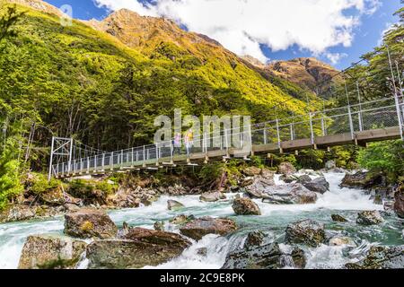 New Zealand tramping people crossing river bridge. Hikers couple backpackers walking hiking together with backpacks on Routeburn Track trail path. Stock Photo