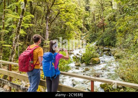 New zealand travel tourists taking phone picture on tramping hike in forest with backpacks. Woman holding smartphone taking photos of river on Stock Photo