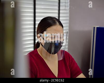 A young and beautiful Asian woman wearing a surgical mask and face shield to protect against Covid-19 while working in an office. Stock Photo