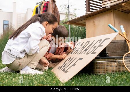 Creative cute kids sitting on grass and making garage sale sign together in backyard Stock Photo