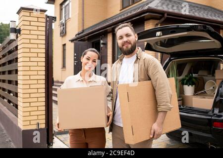Portrait of smiling young couple standing at open car trunk and holding moving boxes against new house Stock Photo