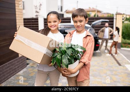 Portrait of positive cute kids holding potted plant and cardboard box in front yard of new house Stock Photo