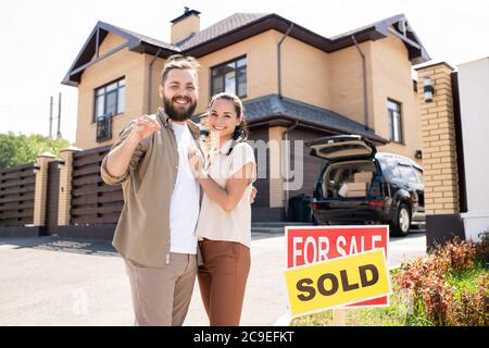 Portrait of a smiling young couple by sold sign standing against sky ...