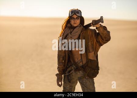 Post-apocalyptic woman with weapon outdoors. Young slim girl warrior in shabby clothes holding sword standing in a confident pose looking at camera . Nuclear post-apocalypse time. Life after doomsday concept. Desert and dead wasteland on the background. Closeup portrait. Stock Photo