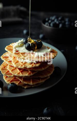 Organic honey is poured on delicious waffles with cheese spread and blueberry fruit, on the grey plate. Stock Photo