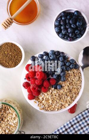 Top down view of fresh muesli breakfast on top of the kitchen table. White bowl with flakes, blueberry, raspberry and honey on white tablecloth. Stock Photo