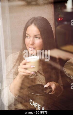 A beautiful girl sits in cafe and looks out window thoughtfully. Reflection of city in window. Brunette woman with long hair drinks cappuccino coffee Stock Photo