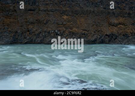 Fast flowing river with dark rocks visible just under the surface. A brown / grey wall of rock rises up on the far side of the river. Quite abstract. Stock Photo