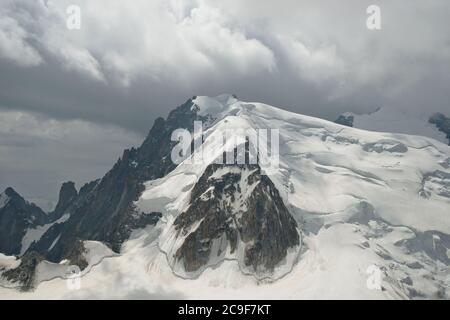 Mountain top with dark rock formations on one side and deep drifts of snow and ice on the other. Bright clouds fill the sky above. Stock Photo