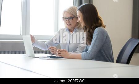 Mature businesswoman teaching female intern, pointing at laptop screen Stock Photo