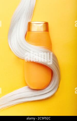 Shampoo bottle wrapped in lock of blonde hair on orange color background. Gold bottle shampoo in dyed hair strand. Hair care cosmetics bath beauty Stock Photo