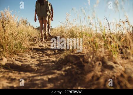 Rear view of two persons walking up the hill on a rocky path. Hikers walking on a rough terrain on a sunny day. Stock Photo