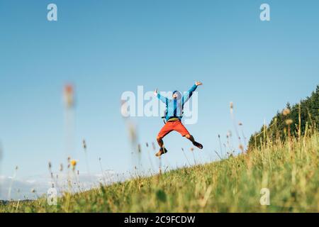 Backpacker traveler emotionally jumping over green grass mountain meadow with backpack with wide opened arms and legs. Human's freedom in nature conce Stock Photo