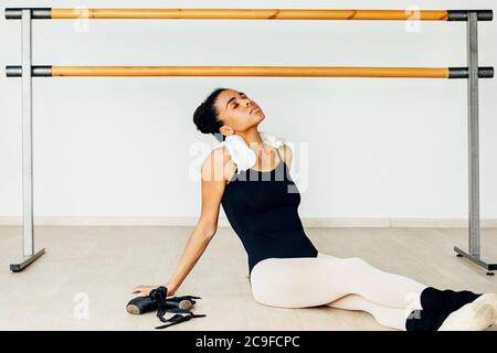 Young ballerina resting after dance training in the studio. Woman with a white towel on her neck resting in dancing class. Stock Photo