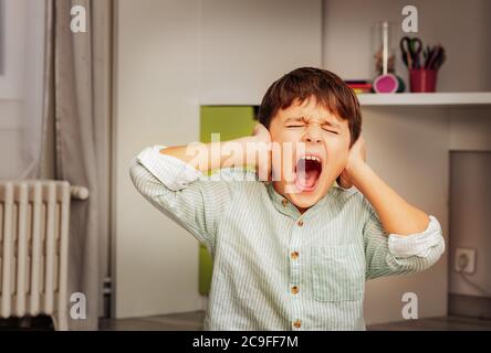 Little autistic boy with strong negative face expression closing ears and loudly screams Stock Photo