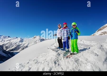 Group of three children stand on the mountain top in snow wearing ski colorful outfit over blue sky Stock Photo